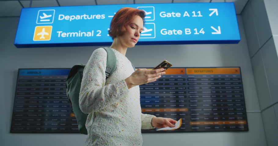 Airport Terminal: Happy Female Passenger Scanning Plane Ticket or Taking Picture Using Phone, Going to Boarding Flight. Traveler Going on Holiday Trip. Digital Flight Information Screen in Background.