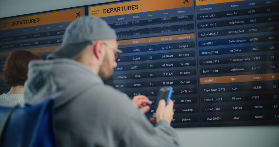 Busy Airport Terminal: Male Passenger with Backpack Looking for Flight on Digital Arrival and Departure Information Board, Using Phone to Check Information for Boarding Plane. Traveler Going on Trip.