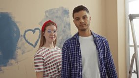 Couple painting new home with blue heart on wall, woman in striped shirt holding brush, smiling indoors, depicting love and creativity in a fresh apartment setting. - Powered by Shutterstock - Get 15% off with code: PIKWIZARD15