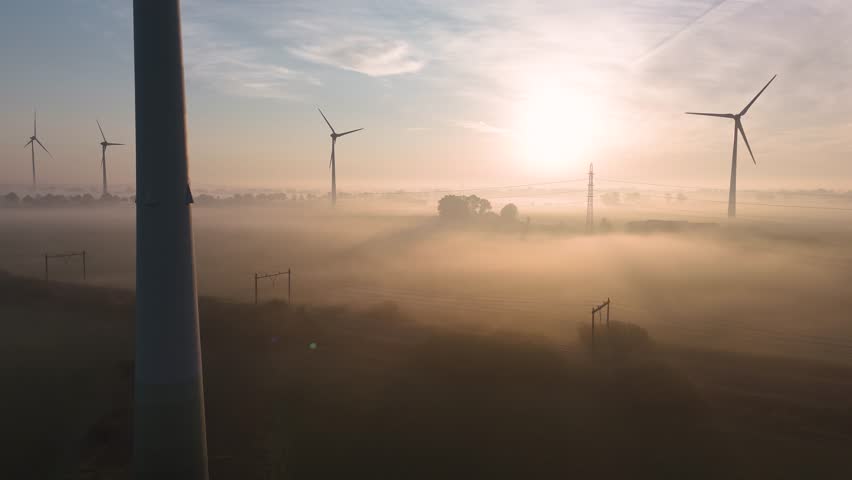 Wind turbines rotate majestically, harnessing wind energy amidst a foggy sunrise over a vast landscape