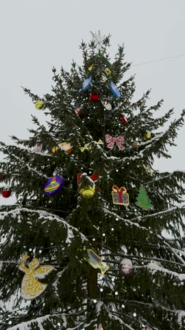 A tall Christmas tree adorned with colorful ornaments, lights, and a star topper. The branches are lightly covered in snow, creating a festive and wintry atmosphere