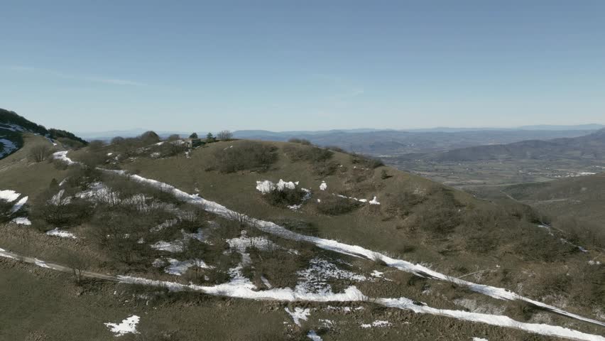Drone view over Valsorda, Gualdo Tadino, on a sunny winter day. Snow-dusted peaks rise above a vibrant landscape, blending crisp white and rich earthy tones under a clear blue sky