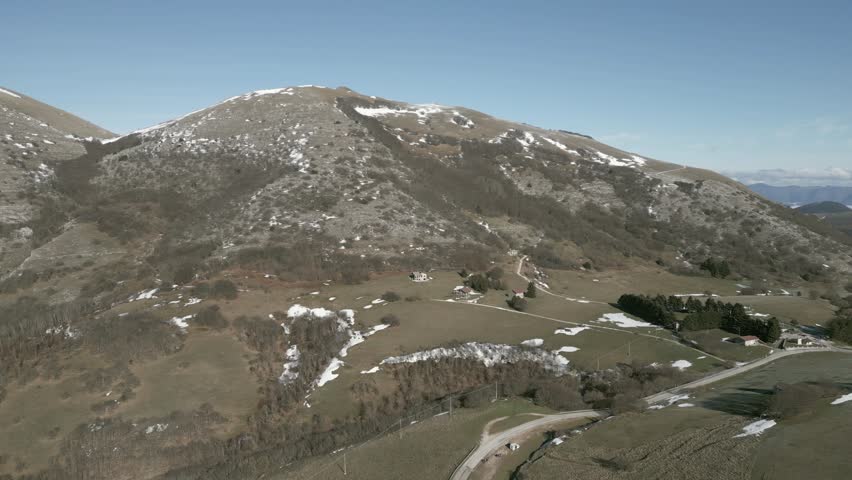 Drone view over Valsorda, Gualdo Tadino, on a sunny winter day. Snow-dusted peaks rise above a vibrant landscape, blending crisp white and rich earthy tones under a clear blue sky