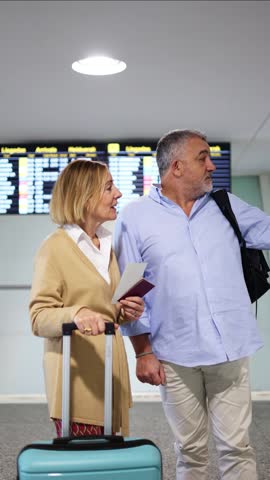 vertical Seasoned senior travelers carefully examining flight schedule on digital screen, holding passports and luggage while standing in bright airport terminal, preparing for international journey
