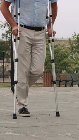 A man barely hobbles on one leg with crutches. Adult male walking slowly down the street, leaning on two crutches. Adult male person walks in the park during the day. Disabled person concept