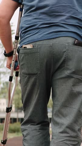 Close-up of man barely hobbles on one leg with crutches. Male in sneakers and gray pants walks slowly down the street, leaning on two crutches. Disabled person walking in park during day. Rear view