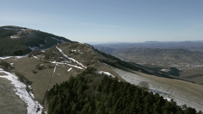 Drone view over Valsorda, Gualdo Tadino, on a sunny winter day. Snow-dusted peaks rise above a vibrant landscape, blending crisp white and rich earthy tones under a clear blue sky