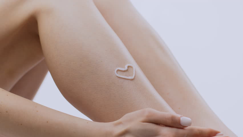 A young woman is pampering herself by applying skin care cream in the shape of a heart on her leg. The backdrop is isolated, allowing focus on her skin care routine and relaxation.
