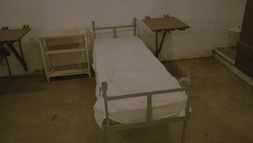 Panning shot of an old sparsely furnished hospital room with simple metal-framed beds, one covered with a thin, coarse brown blanket and the other with white sheet. Hospital of Fort de Mutzig fortress
