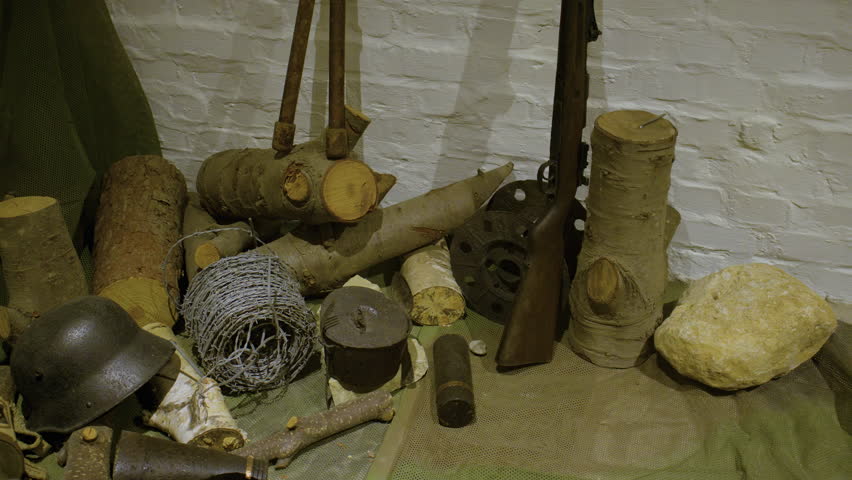 A collection of military artifacts and tools arranged on a green camouflage net. A German World War II helmet, a shovel, and a gas mask rest amongst various pieces of wood