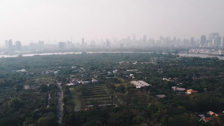 Urban Bangkok landscape revealing verdant fields alongside residential houses, surrounded by towering skyscrapers under hazy atmospheric conditions, showcasing metropolitan environmental contrasts