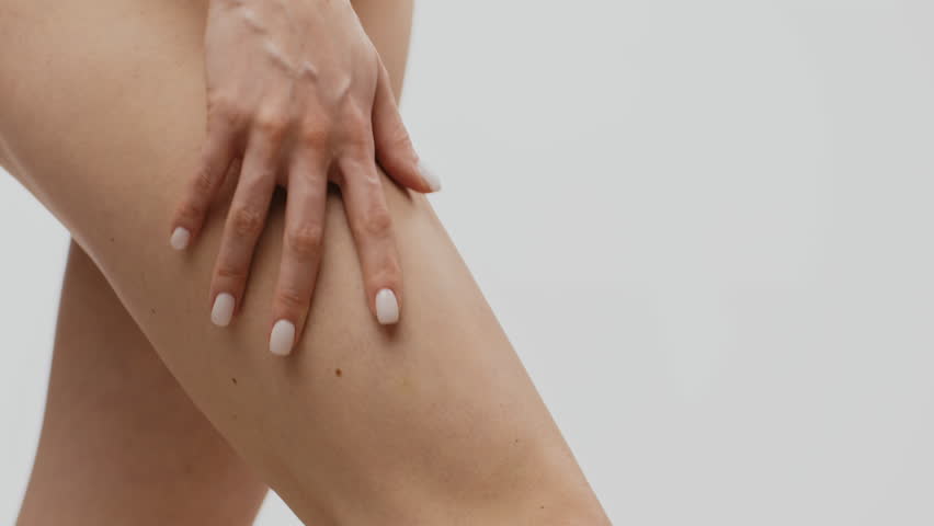 A woman gently applies a nourishing skin care product to her leg, focusing on self-care and pampering. The clean, minimalistic background emphasizes her relaxed atmosphere.