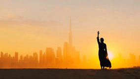 Arabic woman weared in traditional UAE dress - abayain rising her hands on the sunset at a desert with Dubai city silhouette on the background. - Powered by Shutterstock - Get 15% off with code: PIKWIZARD15