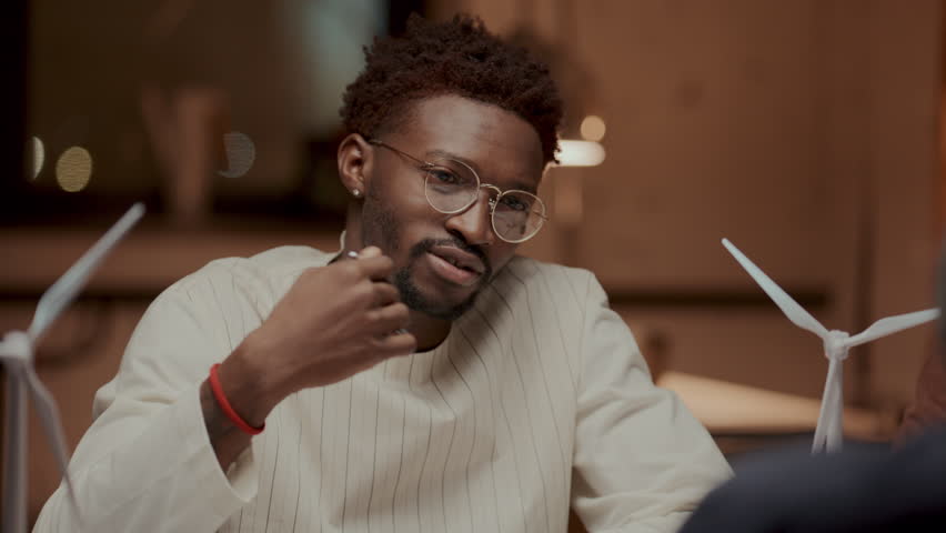 Young Black male engineer in glasses sitting at table with models of wind turbines on it, explaining his ideas for renewable energy project to colleague in office. Over the shoulder view