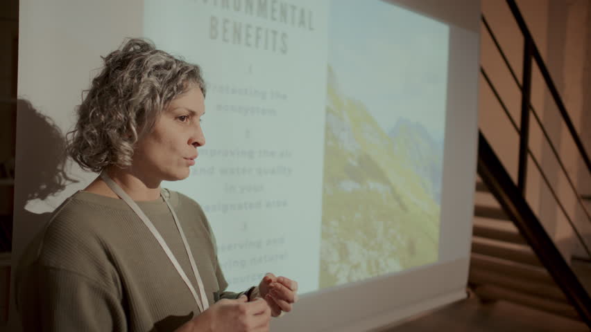 Senior female engineer with curly grey hair giving presentation on energy efficient architecture with slide projected on screen, explaining sustainability concepts to audience in office