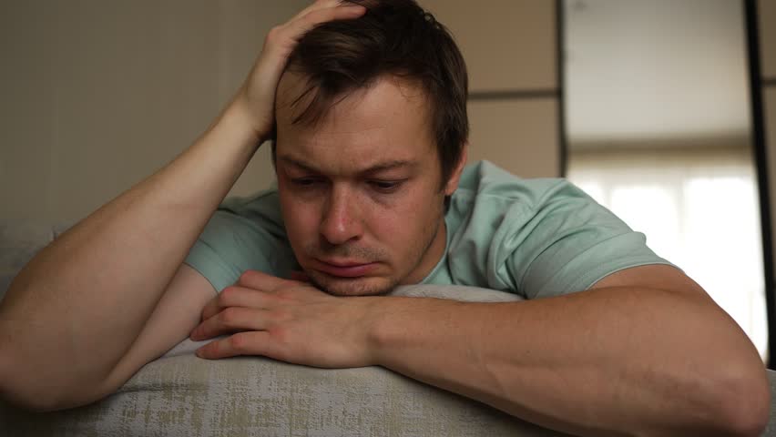 Exhausted young man is lying on sofa at home, holding his head with hand and crying from grief, suffering from severe headache or emotional stress