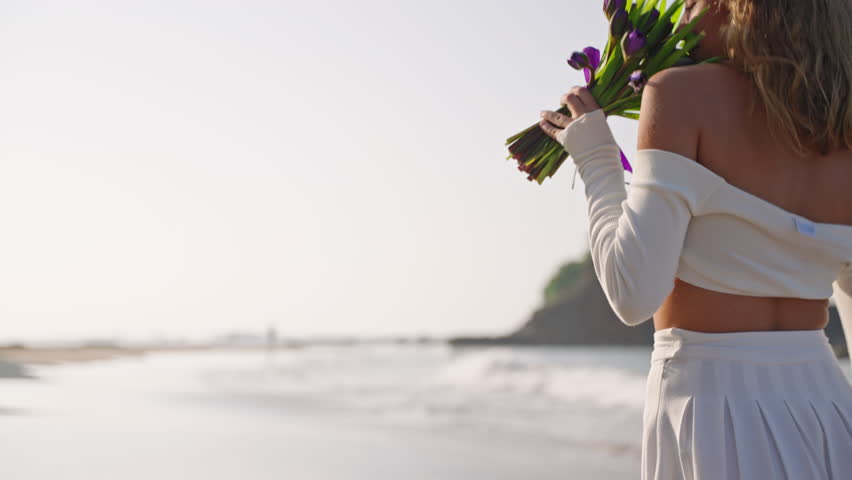 Attractive lady going and smelling bouquet of purple flowers along sandy coast. Female legs walk relaxed on the ocean shore. Woman strolling on seaside at sunset. Close-up, back view.