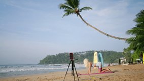 Fitness instructor conducts beach yoga session via live broadcast. Woman demonstrates pose on mat, smartphone and tripod capture online class. Serene tropical backdrop frames digital wellness guide. - Powered by Shutterstock - Get 15% off with code: PIKWIZARD15