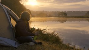 A man with long hair sits by his tent on the shore of a lake during dawn. He pours a hot drink from a thermos, enjoying the silence of nature. The water in the lake reflects the soft golden rays of th - Powered by Shutterstock - Get 15% off with code: PIKWIZARD15