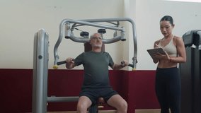 Medium long shot of personal female trainer taking notes on digital tablet while male mature client performing chest press exercise on machine in gym - Powered by Shutterstock - Get 15% off with code: PIKWIZARD15
