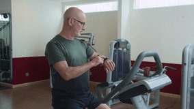 Medium long shot of mature Caucasian man checking his fitness watch while riding exercise bike in gym - Powered by Shutterstock - Get 15% off with code: PIKWIZARD15