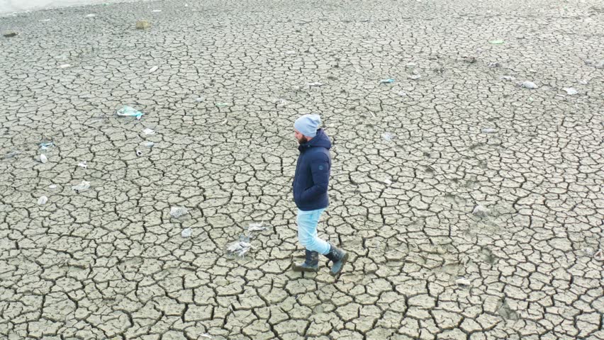 A person walks across a barren landscape showcasing dry, cracked soil caused by drought. The stark environment highlights the effects of climate change.