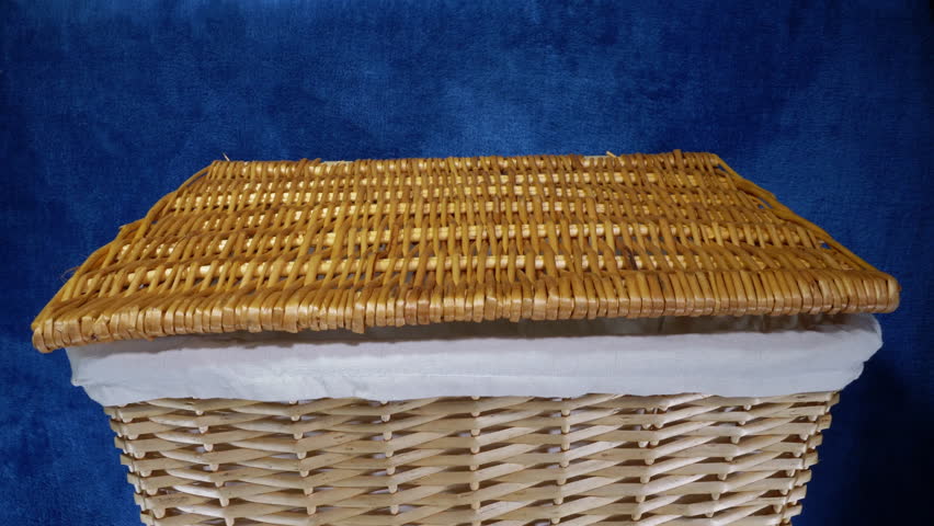 Closeup of a man’s hand opening the lid of a lined wicker laundry basket, then dropping in clothes to be washed and closing the lid.