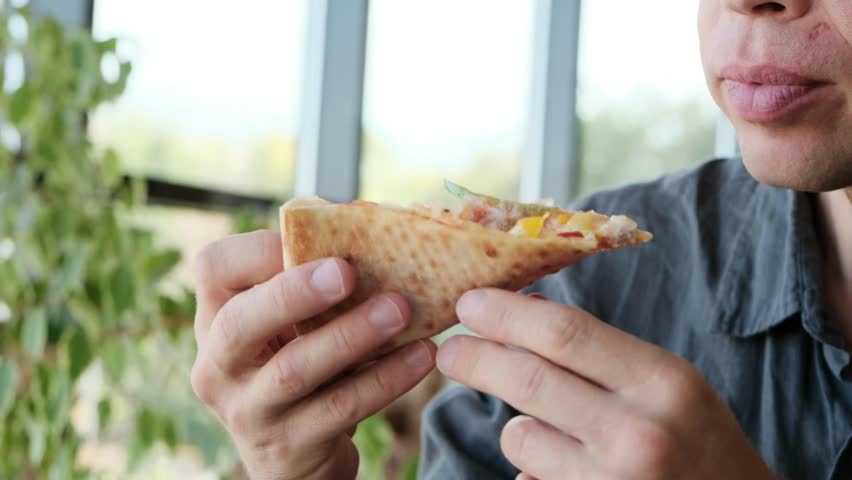 A man in a gray shirt sits at a table in a restaurant, enjoying a slice of pizza. Ideal 4K video for food ads, restaurant promos, and casual dining content.