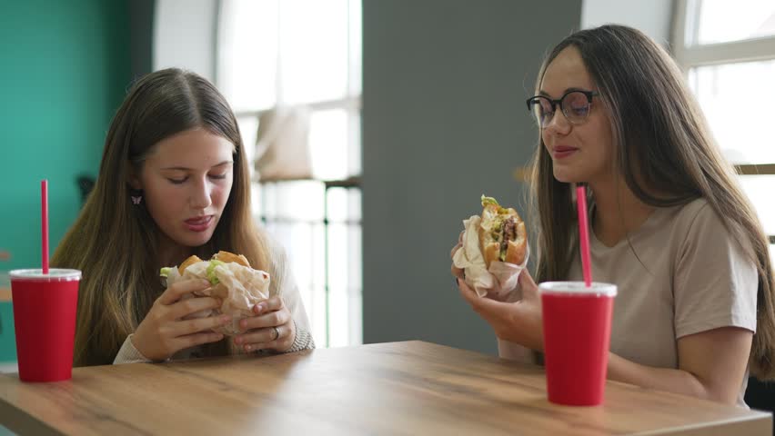 Women relishing food, enjoying sandwiches in vibrant cafe. Casual cafe dining, women savor food, chatting over lunch. Cheerful lunchtime, women with hearty sandwiches in sunlit cafe. Fast food concept