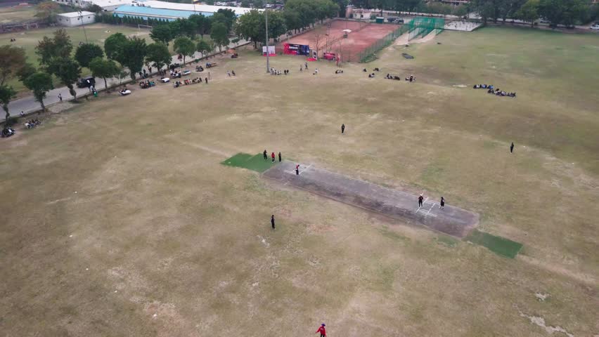 Aerial view of a cricket match in progress on a lush green field. Players are actively engaged, with spectators watching from the sidelines. The scene captures the spirit of outdoor sports and teamwor