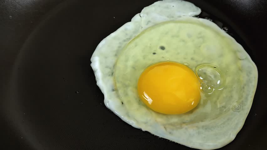 The process of cooking fried eggs in a frying pan directly on a completely black background, horizontal top view