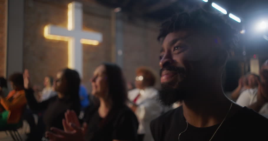 Congregation applauding during a heartfelt worship service, people engaged in faith and prayer, illuminated cross on a brick wall creating a spiritual atmosphere