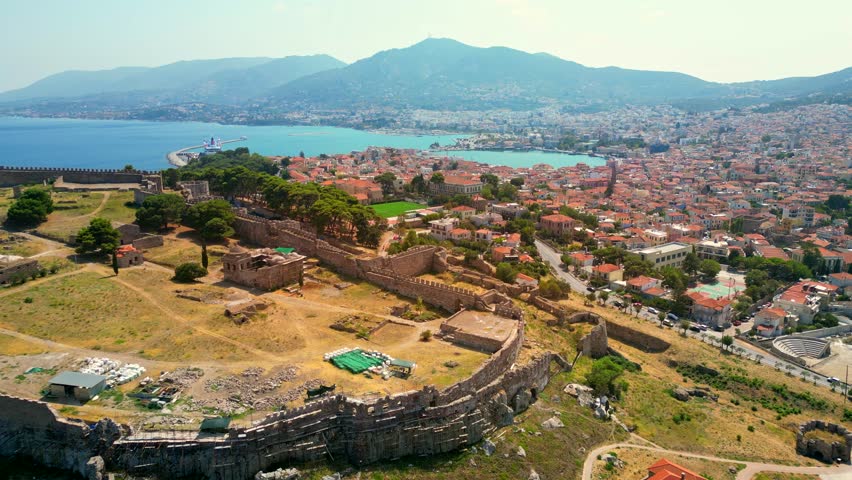 Drone shot passing over the Castle of Mytilene, Greece, overseeing the city and port, with a football pitch, cars, terracotta roofs, and hills in sight.