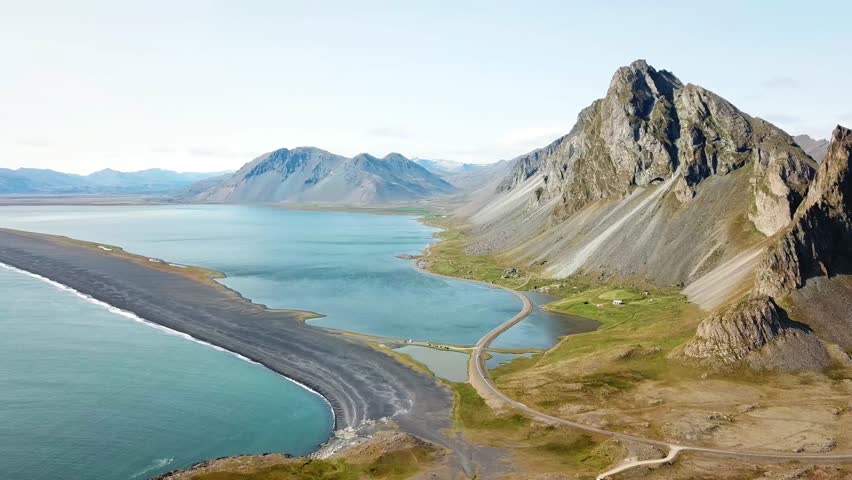 A stunning view of the rugged mountain and serene coastline of Hvalnes, Iceland. The landscape features dramatic cliffs, calm blue waters, and a winding road that blends into the natural beauty