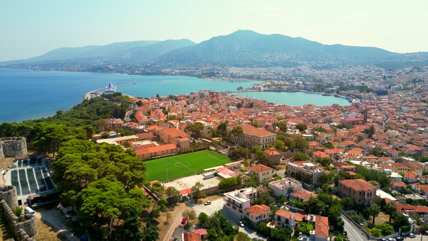 Drone shot orbiting around the city and port of Mytilene, Greece, with a football pitch, traffic, terracotta roofs, and hills in sight.