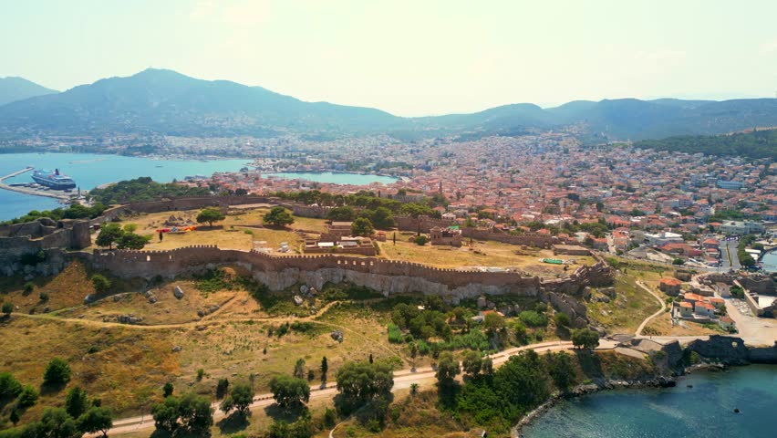 Wide aerial shot orbiting the Castle of Mytilene, Greece, with a backdrop view of the port, terracotta roofed houses, and mountain hills.