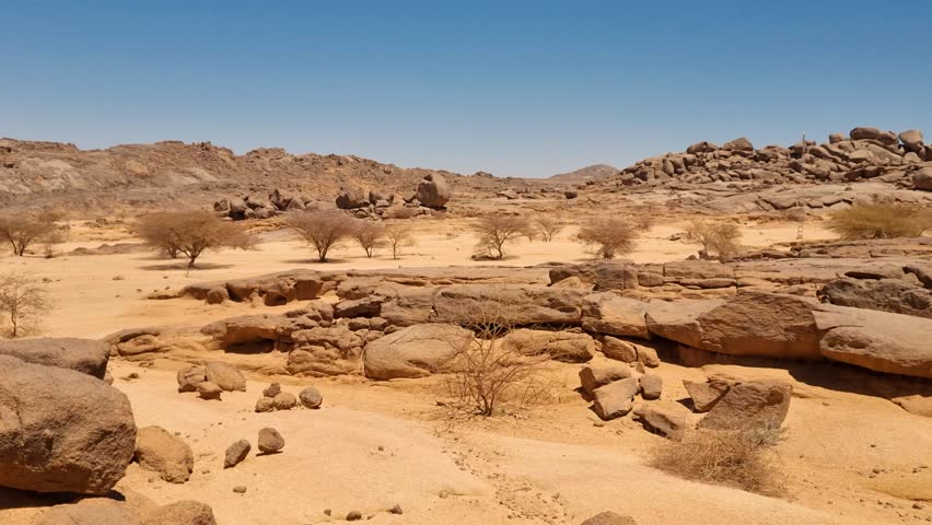 Panning shot of the barren and rugged landscape of the Algerian Sahara Desert on a scorching hot day, rock formations rise amid sparse desert vegetation under the clear blue sky near Tamanrasset
