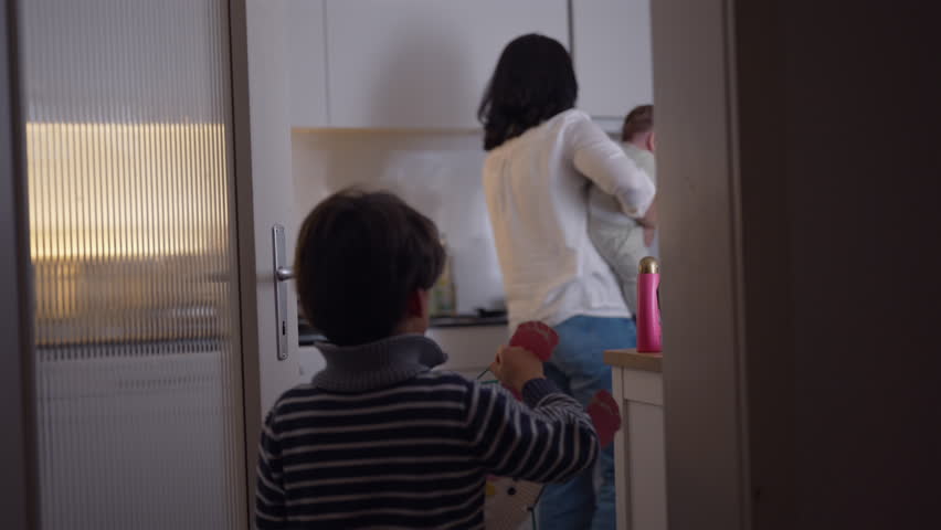 Young boy walking into kitchen holding craft project, mother holding baby near kitchen counter, everyday family interaction during household routine