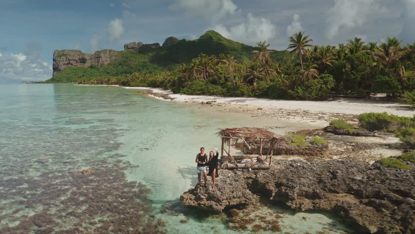 Aerial drone footage captures tourists posing on a rock formation in a beautiful tropical lagoon, with a sandy beach, palm trees, and lush vegetation visible in the background