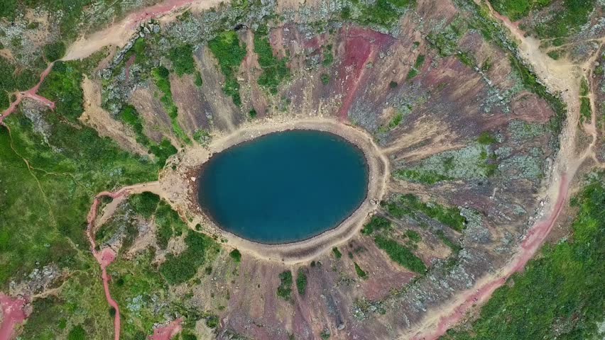 Aerial view of Kerið volcanic crater lake located in Grímsnes area in south Iceland, reflecting blue sky in the Golden Circle, featuring red slopes, green moss and hiking paths, drone top down view