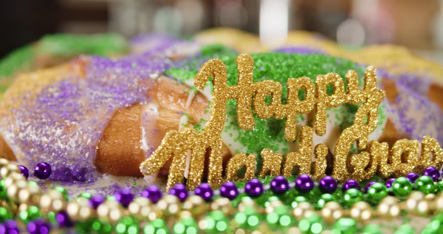 Homemade Mardi Gras king cake with sprinkles in yellow or gold, green, and purple sugar. Surrounded by shiny Mardi Gras beads and glittering golden text, Happy Mardi Gras, to celebrate the tradition.