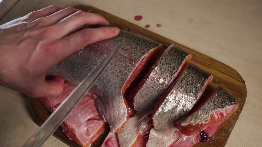 Close-up of chef's hands expertly slicing a fresh salmon fillet into steaks, using a sharp knife on a wooden cutting board, preparing it for cooking