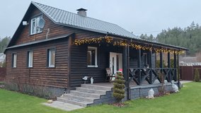 Garlands are lit on the terrace and on a thuja tree growing near the porch of a wooden cottage on Christmas Eve. Nearby grassy lawn, ornamental plants, fence with a gate and forest. Overcast weather - Powered by Shutterstock - Get 15% off with code: PIKWIZARD15