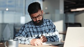 Indian young businessman writes in notebook at modern office desk. Sunlight streams through the window onto the desk. Busy, concentrated worker. - Powered by Shutterstock - Get 15% off with code: PIKWIZARD15