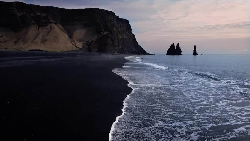 Aerial view of the black sand beach of Reynisfjara, Iceland. Drone moving along the beach with cliffside and rock formations in the distance.