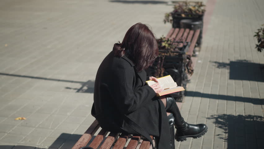 Lady in black coat and leather boots sits on bench in public, leg crossed, reading book, flower pots in background. Urban street with plants and clear sky in the scene