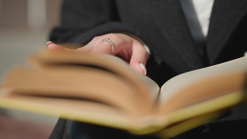 Close-up of hands flipping through book pages, with polished nails adorned with rings, wearing black clothing, background blurred with bokeh effect