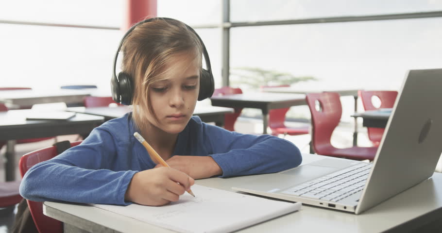 In school, boy wearing headphones writing in notebook, using laptop on desk. Education, learning, studying, technology, student, focus