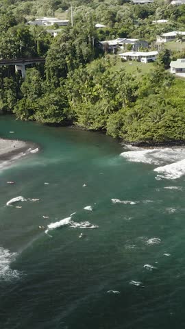 Aerial view of Honolii Beach Park on Hawaii’s Big Island, featuring surfers riding waves, lush tropical greenery, and calm waters.