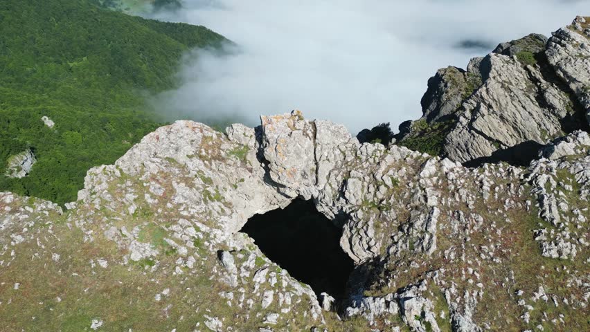 Low clouds are swirling around a rocky mountain ridge with a winding road below on a sunny day
